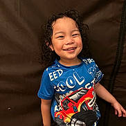 Zackary is registered to the contest to win money with this photo: child, smiling, curly_hair, blue_shirt, monster_truck, red_vehicle, standing, brown_background, happy, toddler, portrait, casual_clothing, indoor, person, cute, joyful, short_sleeves, young, playful, kid