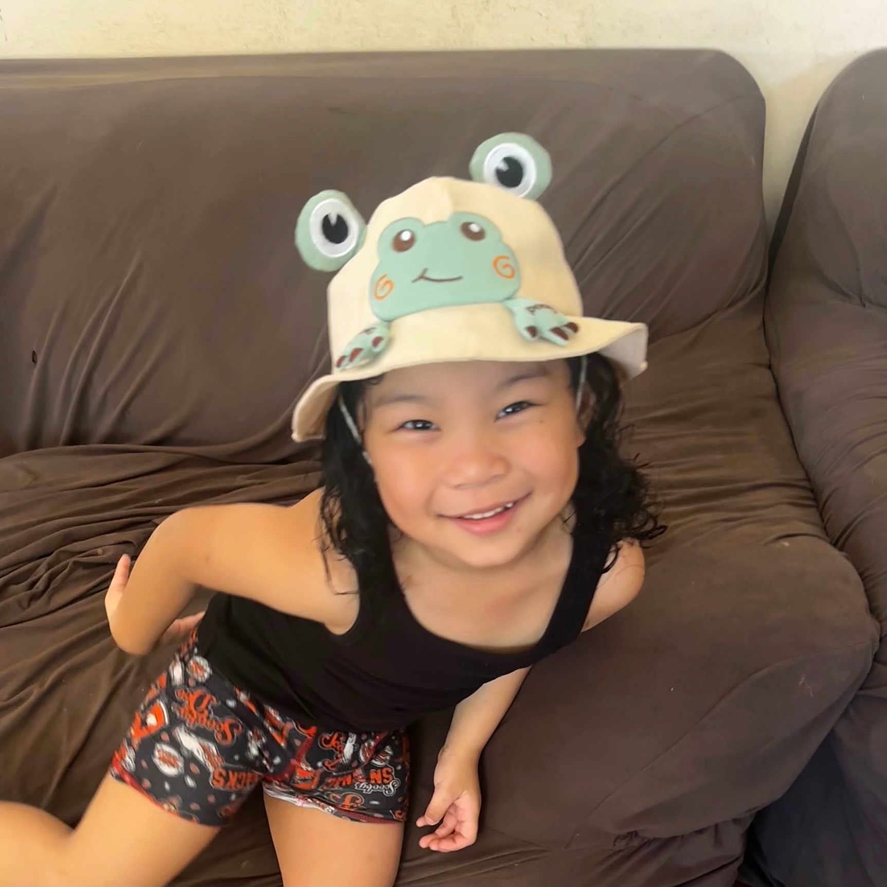 Zackary joined the competition — help win amazing prizes! child, smiling, hat, frog, brown_couch, indoor, black_tank_top, shorts, playful, cute, happy, person, curly_hair, casual, leisure, cozy, home, furniture, portrait, bright