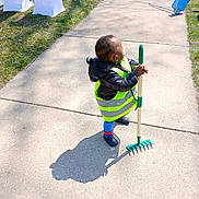 Jamar Leonard joined the competition — help win amazing prizes! child, sidewalk, grass, rake, shadow, jacket, reflective_vest, blue_socks, clogs, outdoor, daylight, sunlight, toy, yard_work, play, person, green, blue, concrete, small