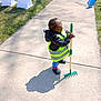 child, sidewalk, grass, rake, shadow, jacket, reflective_vest, blue_socks, clogs, outdoor, daylight, sunlight, toy, yard_work, play, person, green, blue, concrete, small