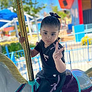 Ainhoa joined the competition — help win amazing prizes! child, girl, carousel, amusement_park, horse, ride, peace_sign, black_clothing, hair_bow, outdoor, daylight, blue_sky, fence, playful, portrait, expression, hand_gesture, fun, youth, person