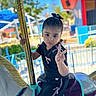 amusement_park, black_clothing, blue_sky, carousel, child, daylight, expression, fence, fun, girl, hair_bow, hand_gesture, horse, outdoor, peace_sign, person, playful, portrait, ride, youth