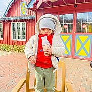 Azlan Muhammad is registered to the contest to win money with this photo: child, bottle, hat, jacket, red_sweater, green_pants, shoes, chair, outdoor, brick_pavement, building, window, door, awning, winter_clothing, cold_weather, curious, serious_expression, daytime, urban