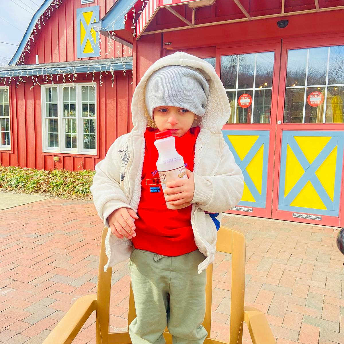 Azlan Muhammad is registered to the contest to win money with this photo: awning, bottle, brick_pavement, building, chair, child, cold_weather, curious, daytime, door, green_pants, hat, jacket, outdoor, red_sweater, serious_expression, shoes, urban, window, winter_clothing