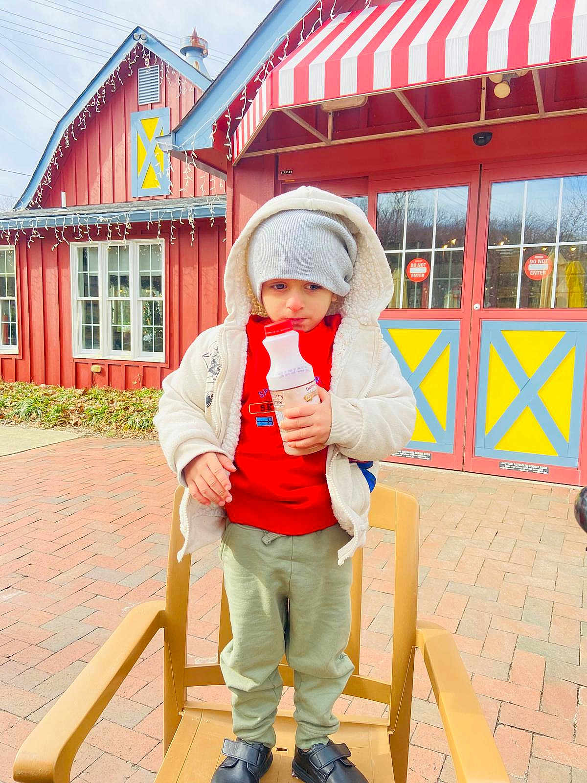 Azlan Muhammad is registered to the contest to win money with this photo: child, bottle, hat, jacket, red_sweater, green_pants, shoes, chair, outdoor, brick_pavement, building, window, door, awning, winter_clothing, cold_weather, curious, serious_expression, daytime, urban