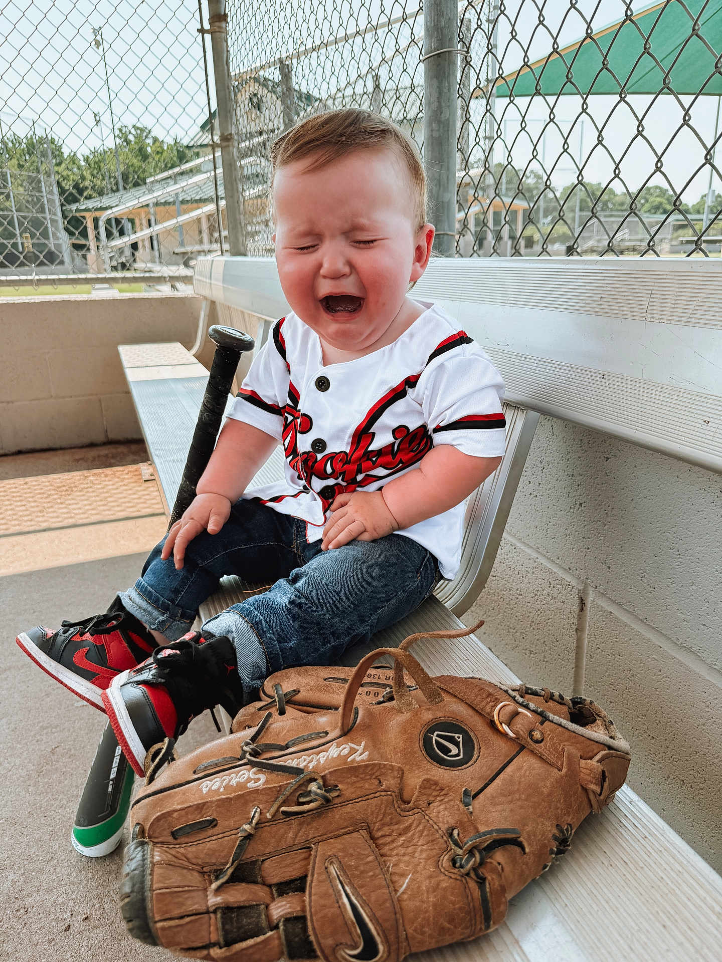 Canaan is registered to the contest to win money with this photo: baby, child, crying, baseball, bench, sports_equipment, glove, bat, fence, outdoor, jeans, sneakers, jersey, person, young_child, sad, park, daylight, metal_bench, recreation