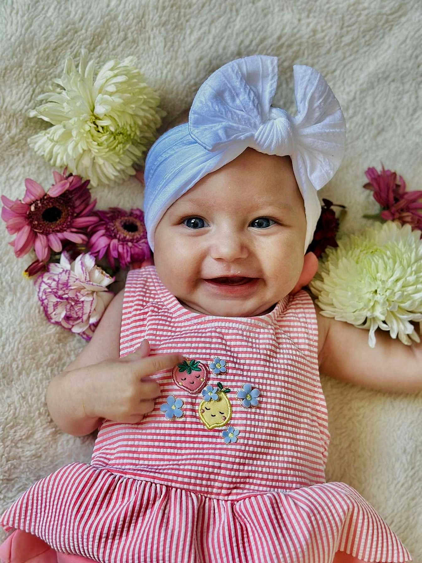 Valentina participe au concours pour gagner de l'argent avec cette photo : baby, child, smiling, headband, bow, flowers, pink_dress, striped_clothing, embroidered_fruits, lying_down, soft_texture, happy, cute, portrait, infant, hand_pointing, blue_eyes, closeup, indoors, colorful