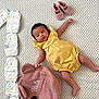 baby, baby_accessories, backpack, child, cute, diapers, footwear, fuzzy, indoor, infant, lying_down, pink, pink_shoes, portrait, question_mark, soft_toys, textured_blanket, white_blanket, yellow, yellow_romper
