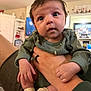baby, infant, close_up, portrait, big_eyes, green_outfit, hands, adult_hand, black_ring, socks, living_room, cabinet, tv, family_photo, cozy, cute, holding, sitting, curious, skin