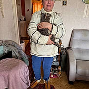 Teddy a rejoint le concours — aidez-le/la à gagner de superbes lots ! person, cat, indoor, sweater, slippers, carpet, armchair, home, living_room, rug, furniture, cozy, pet, gray_cat, portrait, casual, hand, floor, wall, decor