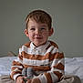 child, boy, smile, striped_shirt, bed, toy, indoor, casual_clothing, cross_legged, young, portrait, happy, cute, cozy, home, sitting, playful, innocent, soft_toy, brown_hair