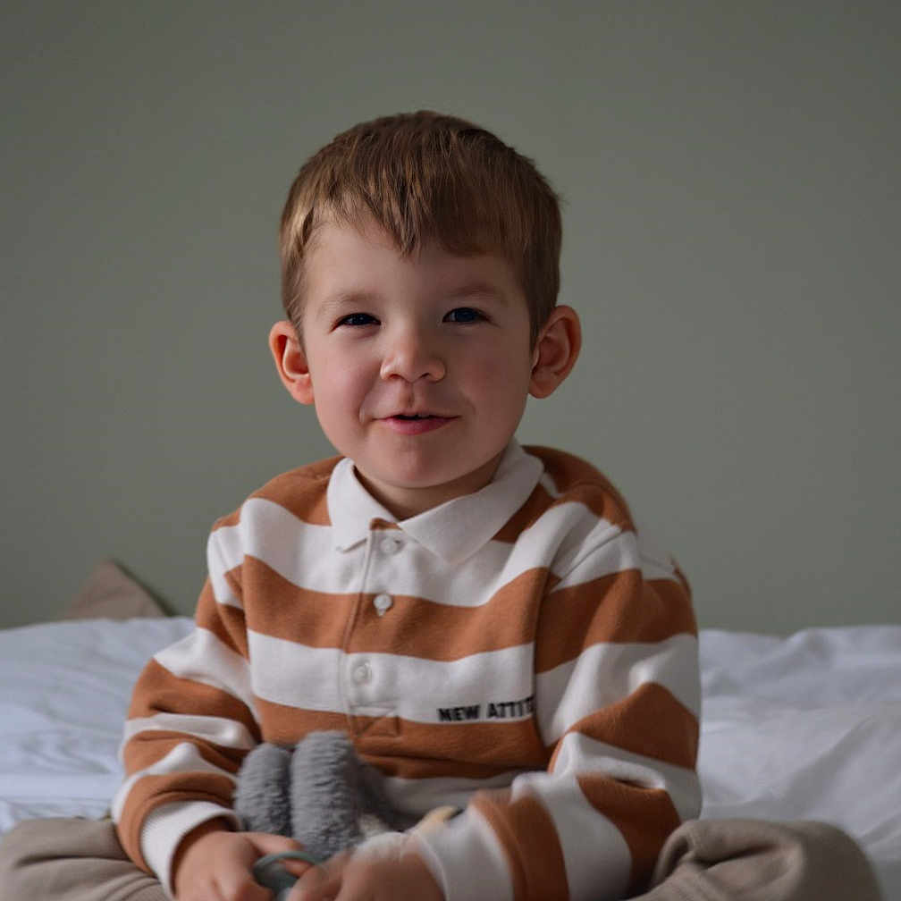 Tomy a rejoint le concours — aidez-le/la à gagner de superbes lots ! bed, boy, brown_hair, casual_clothing, child, cozy, cross_legged, cute, happy, home, indoor, innocent, playful, portrait, sitting, smile, soft_toy, striped_shirt, toy, young