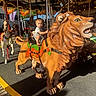child, toddler, carousel, carousel_horse, lion_ride, amusement_park, fairground, outdoor, riding, seated, pole, wooden_figure, animal_figure, playground_ride, colorful, lights, sunlight, portrait, family_outing, entertainment