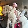 dog, person, indoor, hug, white_dog, pajama_pants, red_plaid, kitchen, counter, water_bottle, headband, light_fixture, cabinet, foot, chair, floor, hand, face, collar, wall_sign