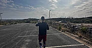 Maximillian is registered to the contest to win money with this photo: asphalt, casual_clothing, clouds, daytime, dog, nature, outdoor, parking_lot, person, plants, rock_wall, shadow, shorts, sky, sneakers, socks, street_lamp, sunny, vehicle, walking