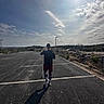 Maximillian is registered to the contest to win money with this photo: person, dog, parking_lot, outdoor, sunny, shadow, asphalt, clouds, sky, walking, casual_clothing, shorts, socks, sneakers, nature, plants, rock_wall, street_lamp, vehicle, daytime