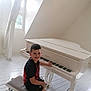 boy, child, piano, white_piano, bench, indoor, smiling, music, musical_instrument, wooden_floor, window, natural_light, short_hair, black_shirt, shorts, footwear, seat, playing, happy, person