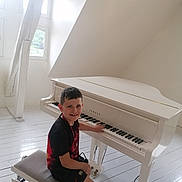 Maël a rejoint le concours — aidez-le/la à gagner de superbes lots ! boy, child, piano, white_piano, bench, indoor, smiling, music, musical_instrument, wooden_floor, window, natural_light, short_hair, black_shirt, shorts, footwear, seat, playing, happy, person