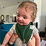 Killian joined the competition — help win amazing prizes! baby, child, indoor, curly_hair, green_bib, person, holding, smiling, cute, living_room, furniture, couch, home_decor, adult_hand, onesie, portrait, infant, happy, close_up, casual