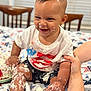Darryn is registered to the contest to win money with this photo: baby, child, smiling, messy, cake, frosting, hands, legs, feet, shirt, shorts, arm, face, home, tablecloth, chair, window, support, celebration, patriotic