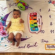 Kinsley is registered to the contest to win money with this photo: baby, infant, yellow_dress, toy_piano, milestone_blanket, flower_pattern, crescent_moon, numbers, star_shape, headband, floor, colorful_toys, curious_expression, indoor, child, playtime, soft_blanket, smiling, cute, lying_down