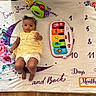 baby, infant, yellow_dress, toy_piano, milestone_blanket, flower_pattern, crescent_moon, numbers, star_shape, headband, floor, colorful_toys, curious_expression, indoor, child, playtime, soft_blanket, smiling, cute, lying_down
