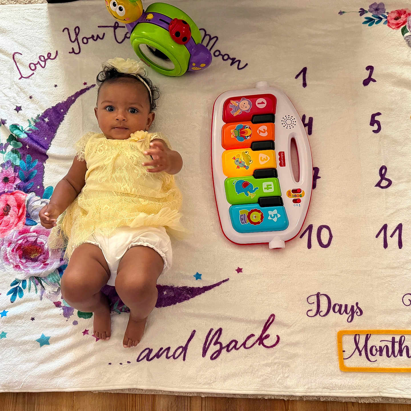 Kinsley is registered to the contest to win money with this photo: baby, infant, yellow_dress, toy_piano, milestone_blanket, flower_pattern, crescent_moon, numbers, star_shape, headband, floor, colorful_toys, curious_expression, indoor, child, playtime, soft_blanket, smiling, cute, lying_down
