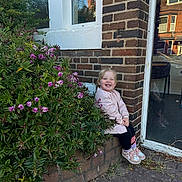 Oliwia joined the competition — help win amazing prizes! child, toddler, smiling, jacket, sneakers, brick_wall, bush, flowers, greenery, outdoor, daylight, window, sidewalk, plant, nature, happy, person, casual_clothing, building, reflection