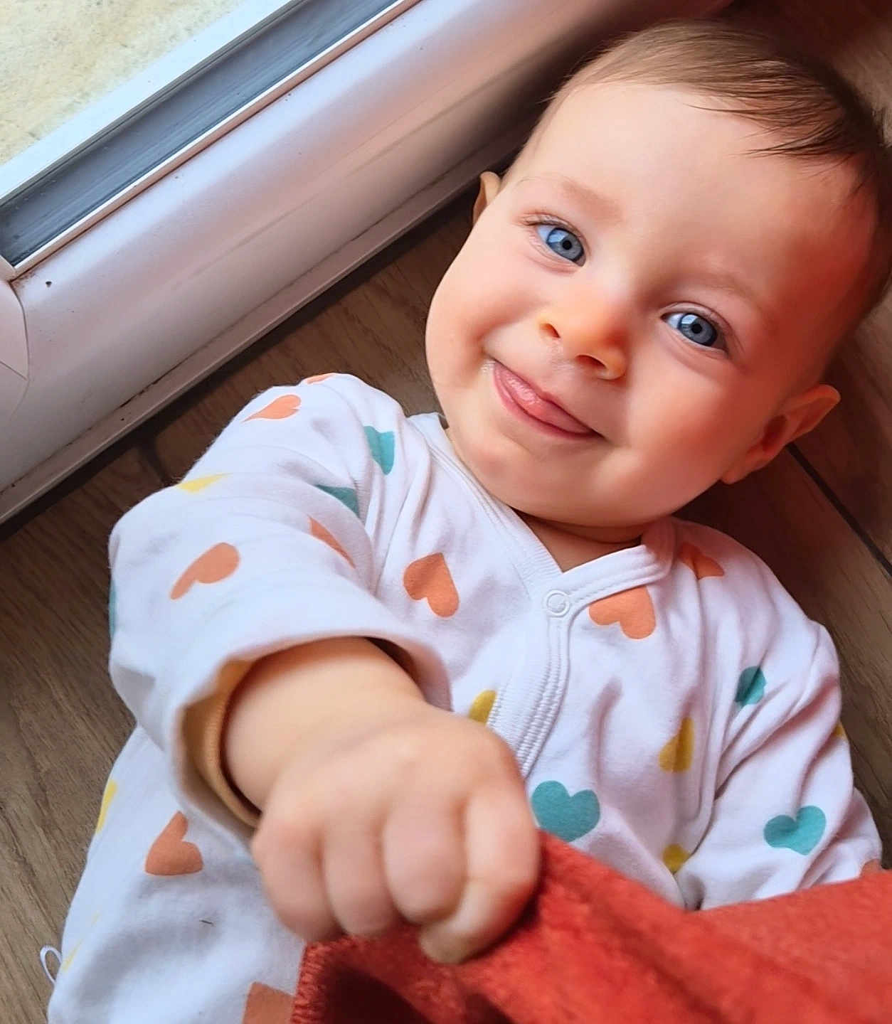 Athèna participe au concours pour gagner de l'argent avec cette photo : baby, infant, child, blue_eyes, smile, onesie, heart_pattern, hand, floor, window, natural_light, playful, face, cute, indoors, portrait, skin, young_child, happy, clothing