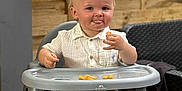 Sébastien participe au concours pour gagner de l'argent avec cette photo : toddler, child, high_chair, food, white_shirt, brown_pants, yellow_socks, indoor, wooden_wall, table, furniture, face, smiling, playful, person, cute, baby, seat, tongue_out, eating