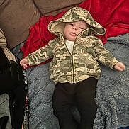 Ezra is registered to the contest to win money with this photo: baby, child, hoodie, camouflage, blanket, red_blanket, blue_blanket, boots, pants, clothing, face, person, infant, couch, indoor, floor, cozy, lying_down, casual, cute