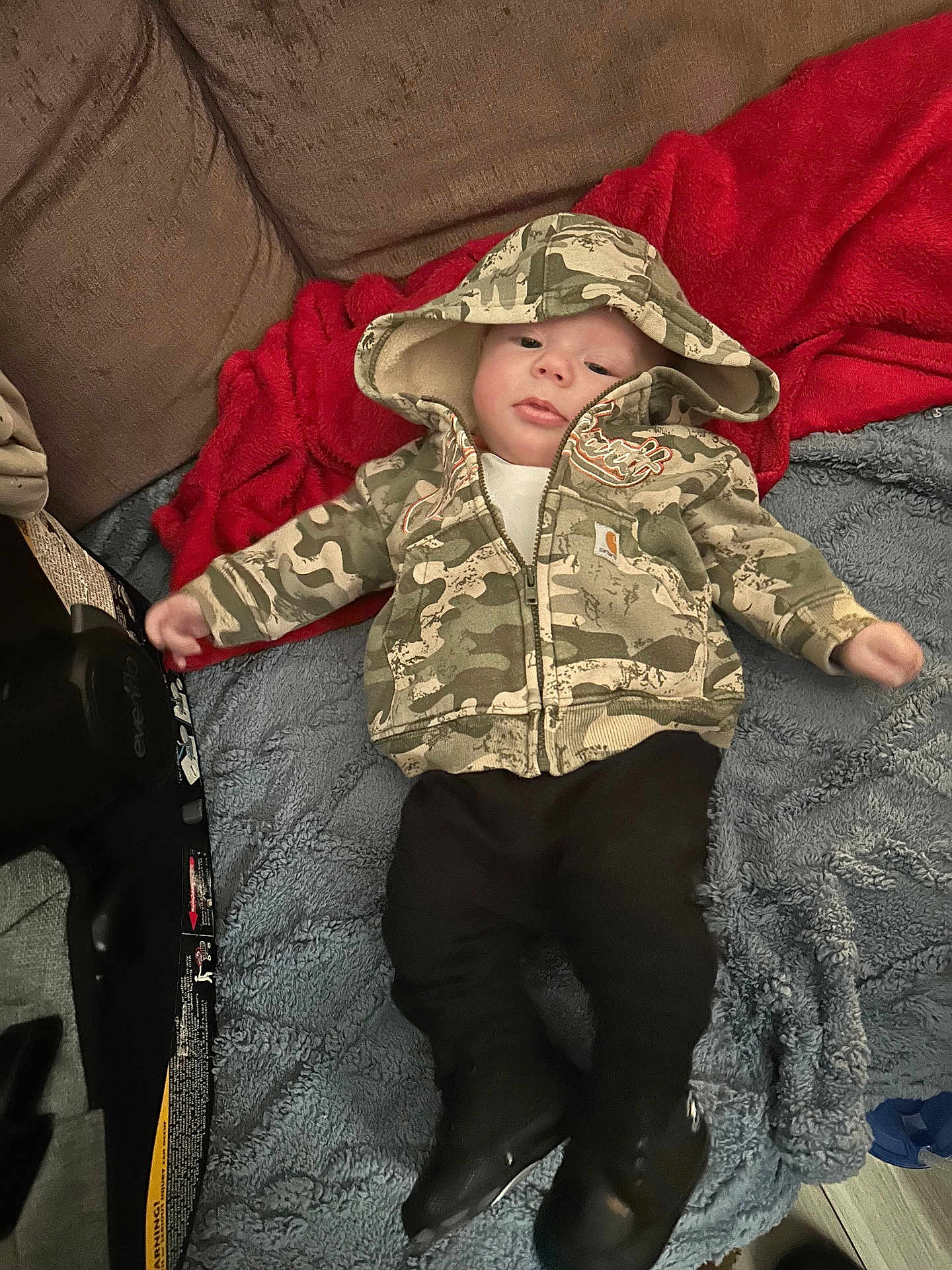 Ezra is registered to the contest to win money with this photo: baby, child, hoodie, camouflage, blanket, red_blanket, blue_blanket, boots, pants, clothing, face, person, infant, couch, indoor, floor, cozy, lying_down, casual, cute