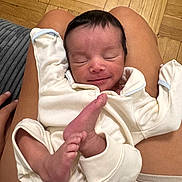 Auralise is registered to the contest to win money with this photo: newborn, baby, infant, sleeping, smiling, portrait, onesie, white_clothing, bare_feet, feet, crossed_legs, cozy, indoors, wooden_floor, adult_lap, closeup, peaceful, family, lying_down, photography