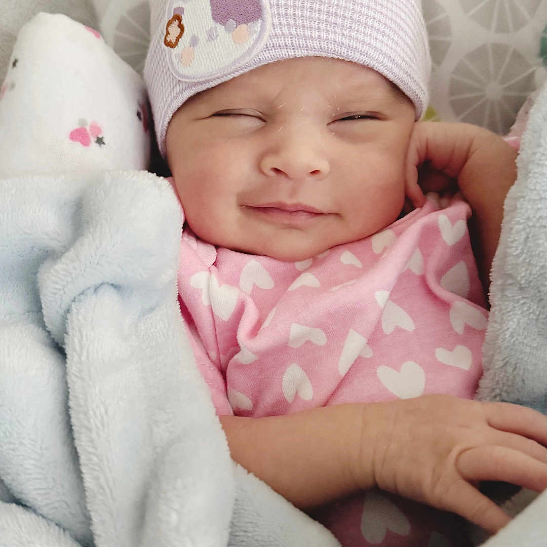 Norrakkesa is registered to the contest to win money with this photo: baby, blanket, child, cozy, cute, cute_expression, face, hat, headwear, heart_pattern, indoors, infant, newborn, pink_clothing, resting, sleepy, smiling, soft_texture, warm, wrapped