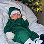 baby, sleeping, blanket, christmas_tree, ornaments, lights, green_outfit, nike, headband, socks, footwear, indoor, holiday, festive, cozy, child, infant, soft_texture, decorations, resting