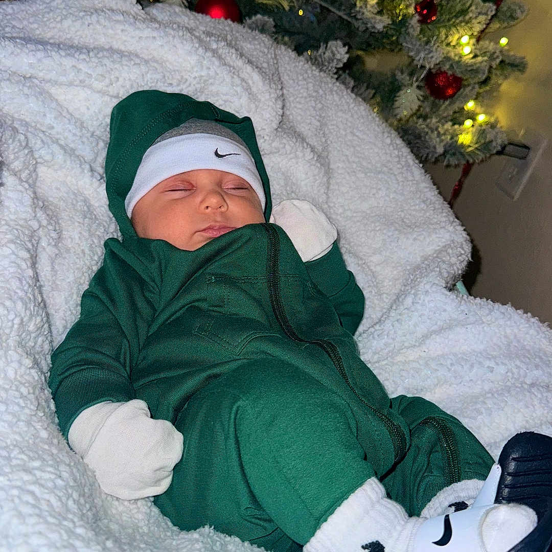 𝘓𝘢𝘩𝘪𝘢𝘮 is registered to the contest to win money with this photo: baby, blanket, child, christmas_tree, cozy, decorations, festive, footwear, green_outfit, headband, holiday, indoor, infant, lights, nike, ornaments, resting, sleeping, socks, soft_texture