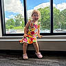 toddler, child, smiling, portrait, indoors, window, windowsill, dress, colorful_clothing, fruit_pattern, pink_shoes, clogs, carpet, toy, sunlight, blue_sky, clouds, trees, parking_lot, happy