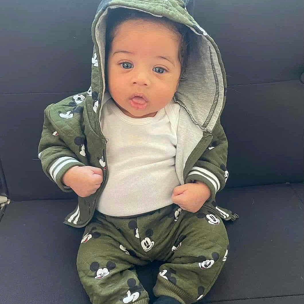 Jaylen is registered to the contest to win money with this photo: baby, child, hoodie, green_clothing, cartoon_print, pants, sitting, couch, indoor, cute, curious, infant, face, portrait, clothing, fists, expression, young_child, socks, casual_wear