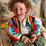 Valentin a rejoint le concours — aidez-le/la à gagner de superbes lots ! child, toddler, curly_hair, smiling, colorful_jacket, striped_jacket, green_pants, checkered_pants, sitting, indoor, nursery, changing_table, wall_art, elephant_mural, teddy_bear_mural, happy, playful, person, clothing, footwear