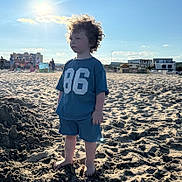 Valentin a rejoint le concours — aidez-le/la à gagner de superbes lots ! child, toddler, beach, sand, sunlight, curly_hair, blue_clothing, barefoot, outdoor, summer, sky, clouds, people_in_background, buildings, casual_wear, daytime, portrait, standing, number_86, sun