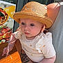adult_hand, baby, child, closeup, curious_expression, food_poster, glass_door, hat, holding, infant, light_clothing, outdoor, patterned_clothing, person, portrait, red_nail_polish, straw_hat, sunlight, table, tattoo