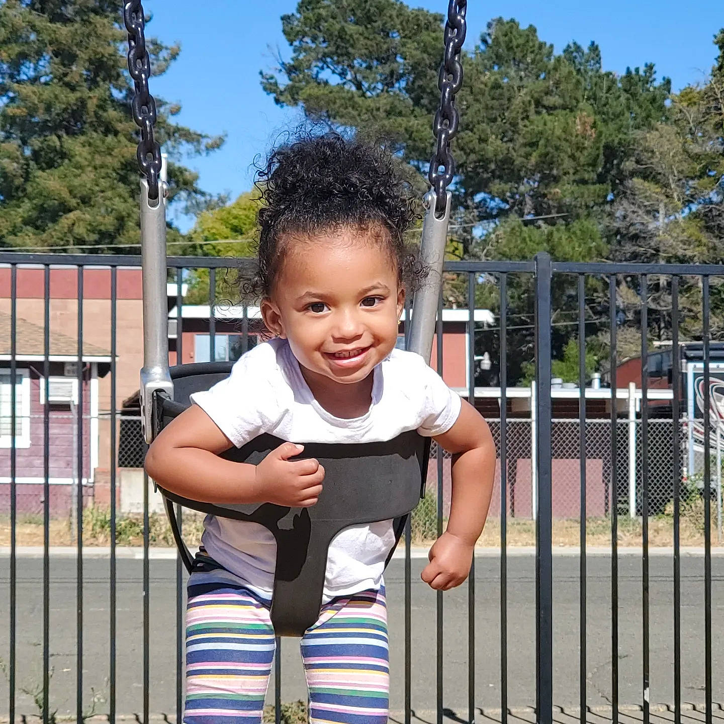 Mia is registered to the contest to win money with this photo: baby, baby_toddler_clothing, child, city, fence, fun, grass, happy, joy, leisure, outdoor_play_equipment, people_in_nature, person, plant, play, recreation, sky, sleeve, smile, t_shirt