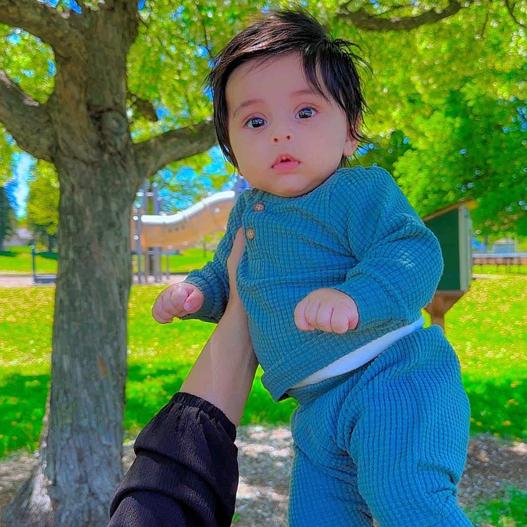 Yaman is registered to the contest to win money with this photo: baby, child, person, outdoor, park, tree, grass, sunlight, hand, clothing, blue_pajamas, face, playing, nature, daylight, greenery, holding, cute, young_child, expression