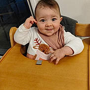 Leïla a rejoint le concours — aidez-le/la à gagner de superbes lots ! baby, child, high_chair, wooden_furniture, indoor, bib, reindeer_design, curly_hair, cute, smiling, playful, clothing, pink, white, person, face, table, seat, hand, expression