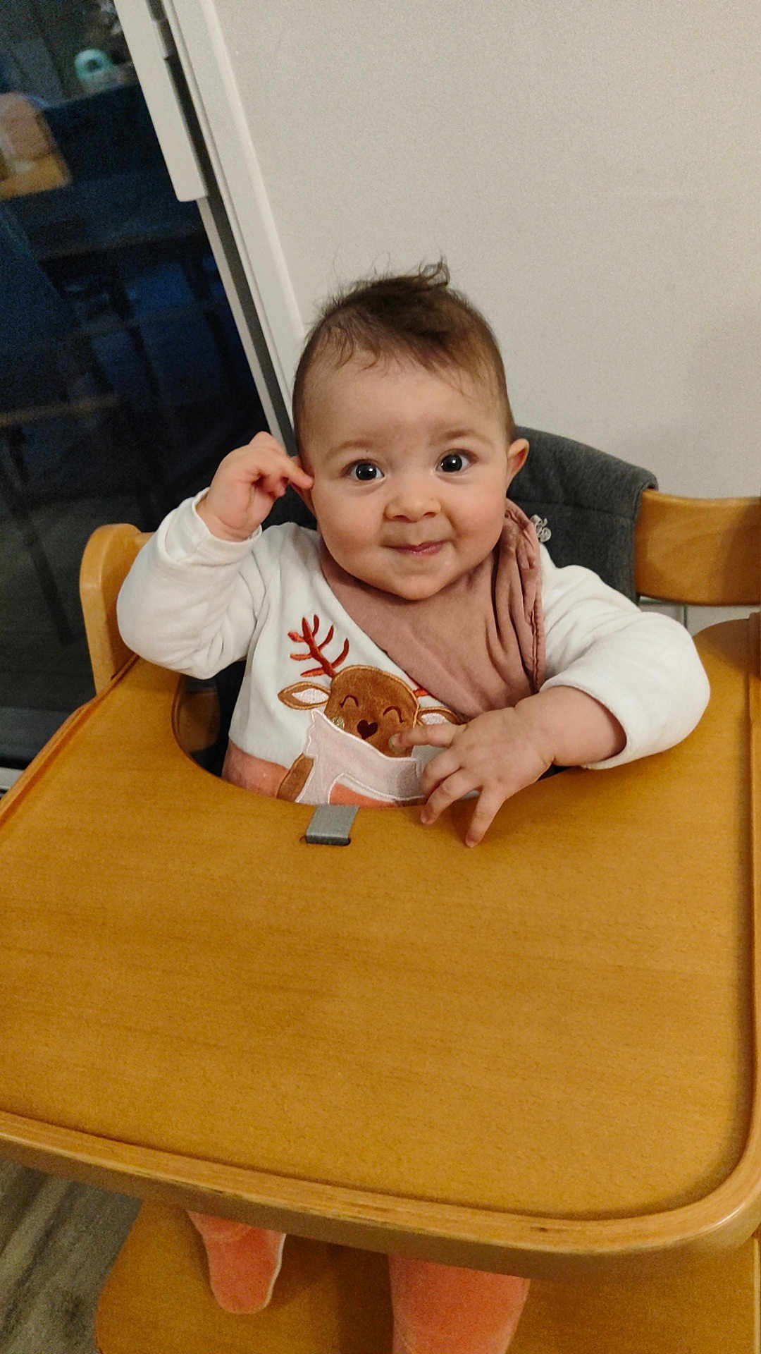 Leïla a rejoint le concours — aidez-le/la à gagner de superbes lots ! baby, child, high_chair, wooden_furniture, indoor, bib, reindeer_design, curly_hair, cute, smiling, playful, clothing, pink, white, person, face, table, seat, hand, expression