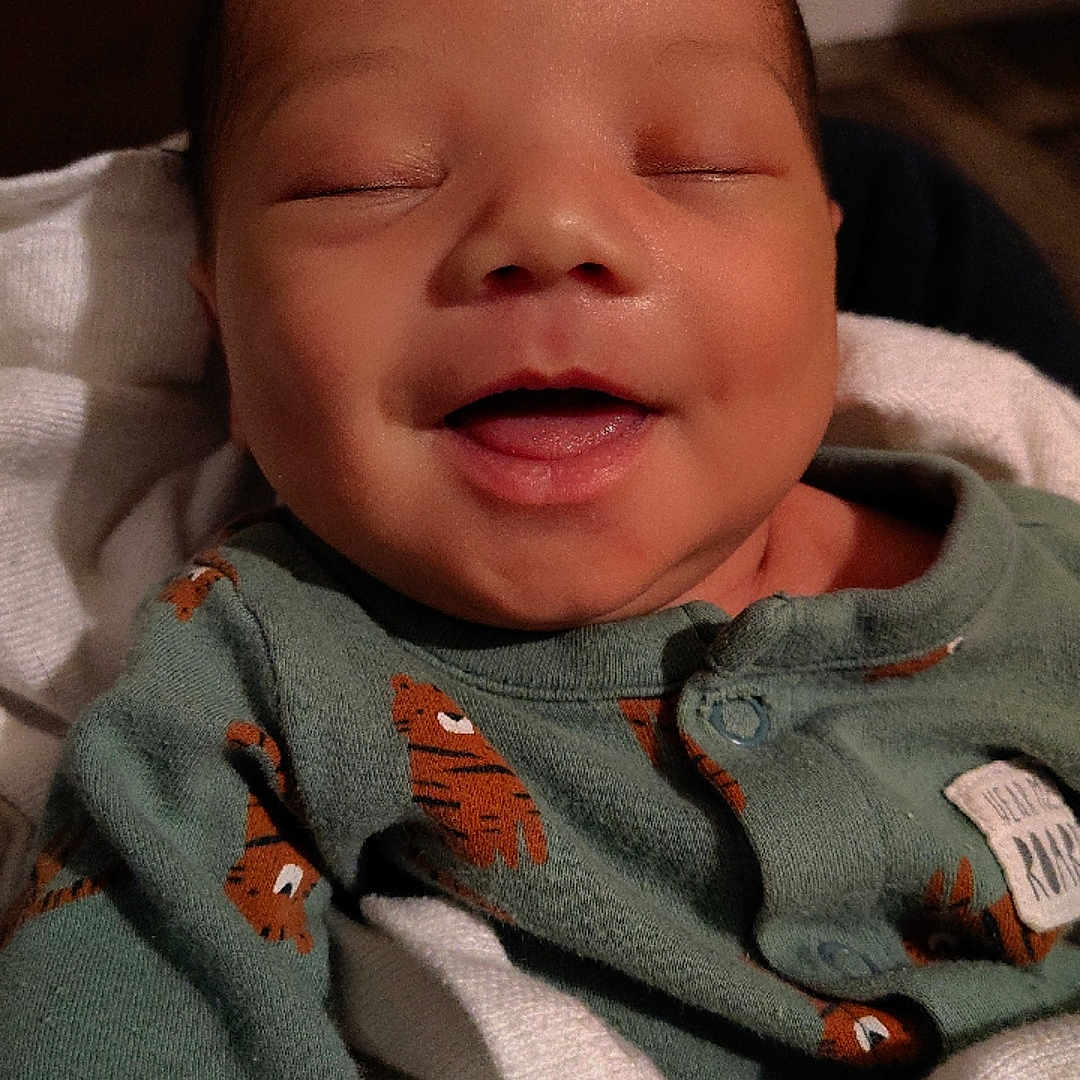 Wynston is registered to the contest to win money with this photo: baby, blanket, child, close_up, clothing, cozy, cute, face, green_onesie, happy, human, indoors, infant, newborn, peaceful, portrait, sleepy, smiling, soft_lighting, tiger_print