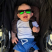 Lincoln joined the competition — help win amazing prizes! baby, child, sunglasses, stroller, smiling, happy, white_shirt, blue_shorts, drawstring, seatbelt, reflection, outdoor, sunlight, infant, person, cute, casual_clothing, headwear, seat, portrait