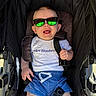 baby, child, sunglasses, stroller, smiling, happy, white_shirt, blue_shorts, drawstring, seatbelt, reflection, outdoor, sunlight, infant, person, cute, casual_clothing, headwear, seat, portrait