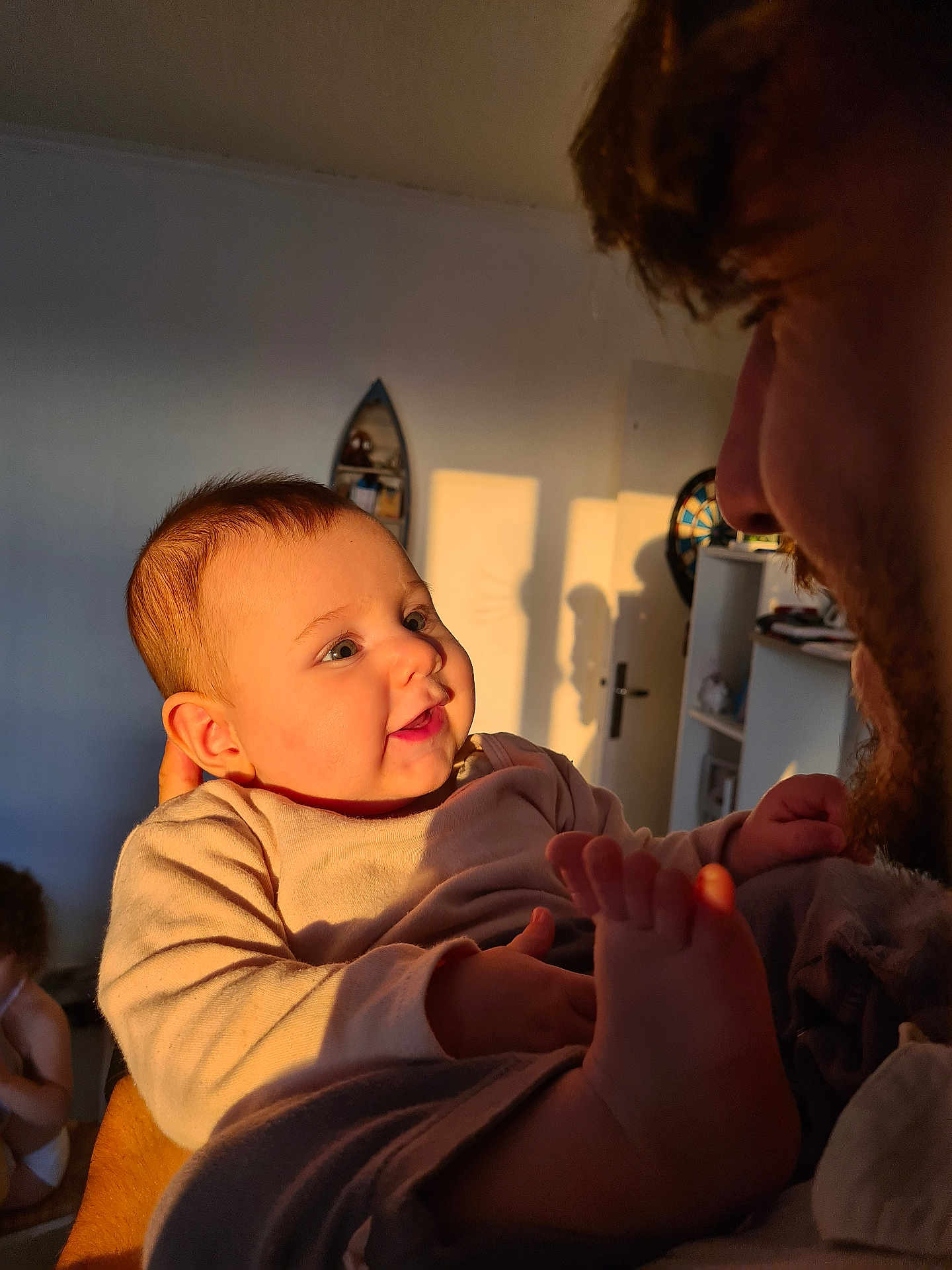 Amalïa a rejoint le concours — aidez-le/la à gagner de superbes lots ! adult, baby, beard, close_up, cozy, door, face, foot, furniture, hand, indoor, infant, interaction, parent_child, person, shadow, shelf, smile, sunlight, warm_light