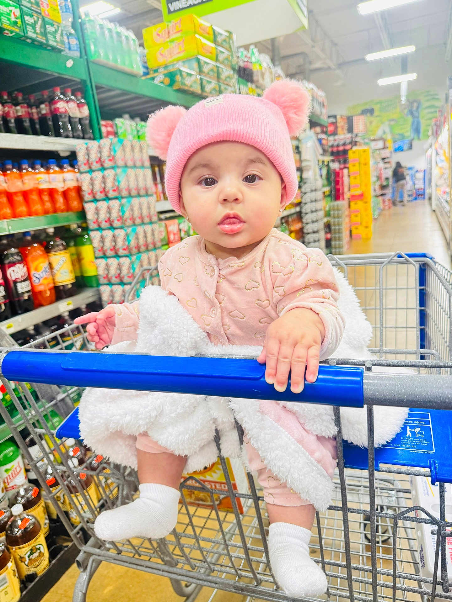 Eimmy joined the competition — help win amazing prizes! baby, child, shopping_cart, grocery_store, pink_hat, pom_poms, blanket, socks, curious, shelf, beverages, soda, shopping, retail, infant, cute, indoor, clothing, person, cart_handle